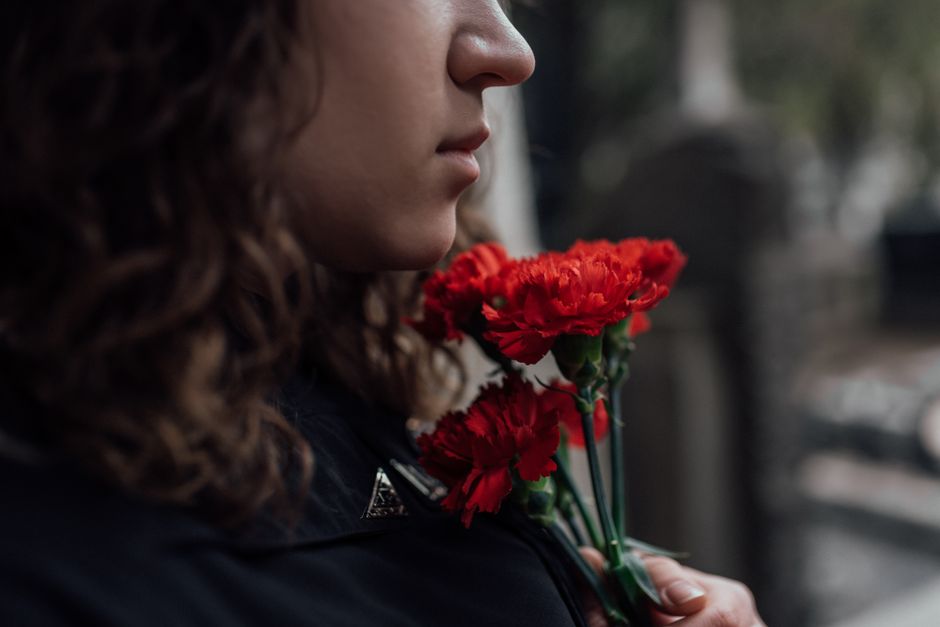 A woman with curly brown hair holding a small bouquet of bright red carnations close to her face, in a contemplative moment outdoors. She is wearing a dark jacket, and the flowers are wrapped in clear