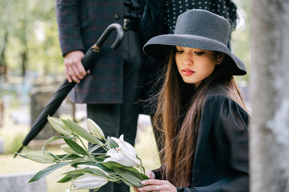 A young woman with long brown hair wearing a wide-brimmed navy hat and black coat holds a bouquet of white lilies with green leaves, wrapped in a simple clear plastic covering. She gazes softly at the