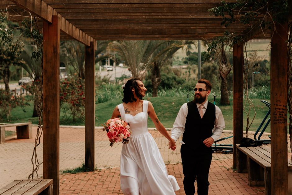 A bride and groom walk hand in hand beneath a wooden pergola in an outdoor park setting. The bride wears a white, sleeveless, A-line wedding dress with a fitted bodice, and holds a bouquet of pink and