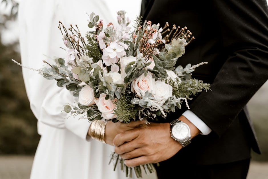 A close-up of a couple holding a floral bouquet during a wedding, featuring soft pastel pink roses, white blooms, and green foliage with hints of eucalyptus and textured branches, arranged in a loose,