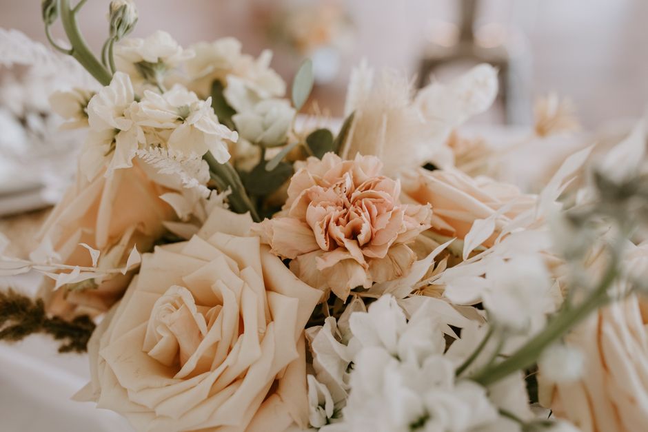 A floral arrangement featuring soft peach and ivory roses, white lisianthus, and dried pampas grass, all arranged in a loose, natural style. The bouquet is set on a light-colored surface with natural 