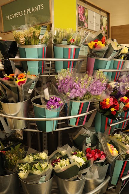 An indoor display of various fresh floral bouquets arranged in metal buckets and striped paper wraps on a multi-tiered rotating stand. The flowers include lilac, yellow, pink, red, and white blossoms 