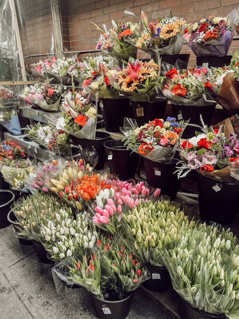 A collection of fresh floral bouquets arranged in black buckets, featuring a variety of vibrant flowers including pink and white tulips, orange carnations, and mixed seasonal blooms. The bouquets are 